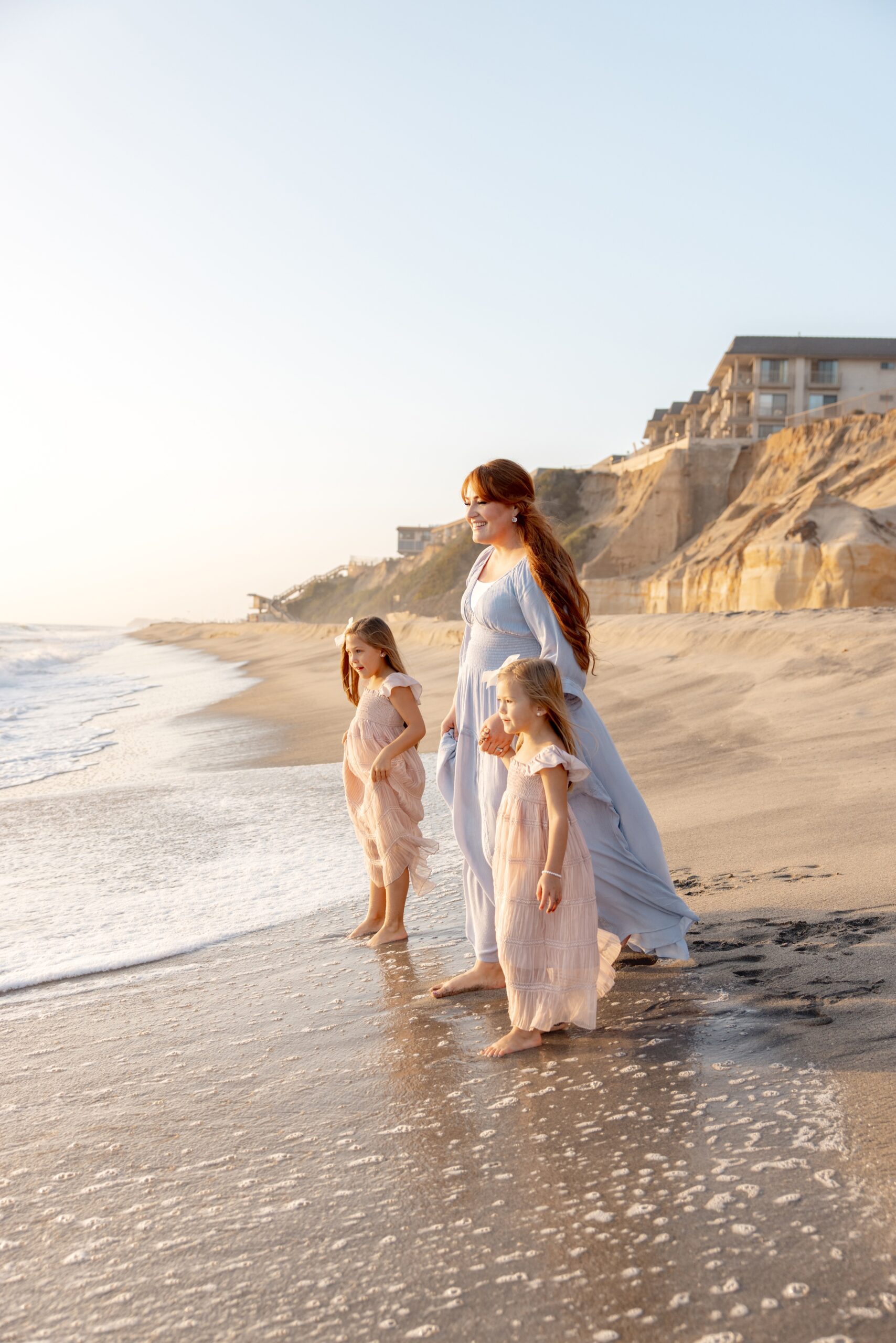 A mom in a blue dress walks her toddler daughters on the beach at sunset after riding a hot air balloon in san diego