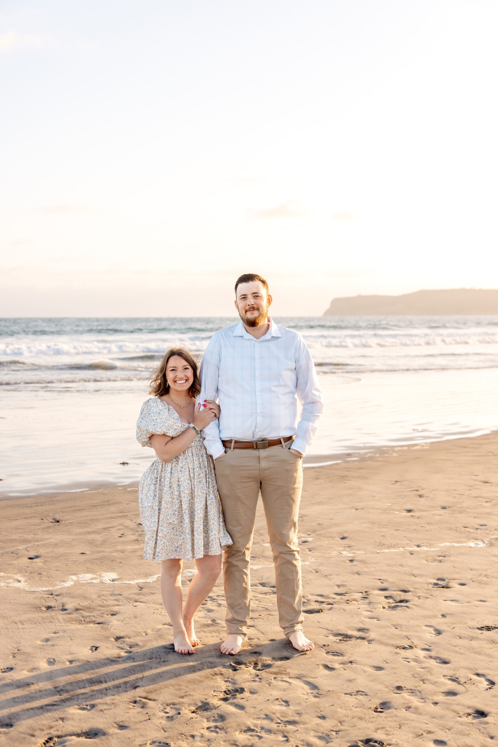 A man and his wife stand on a beach smiling and cuddling at sunset