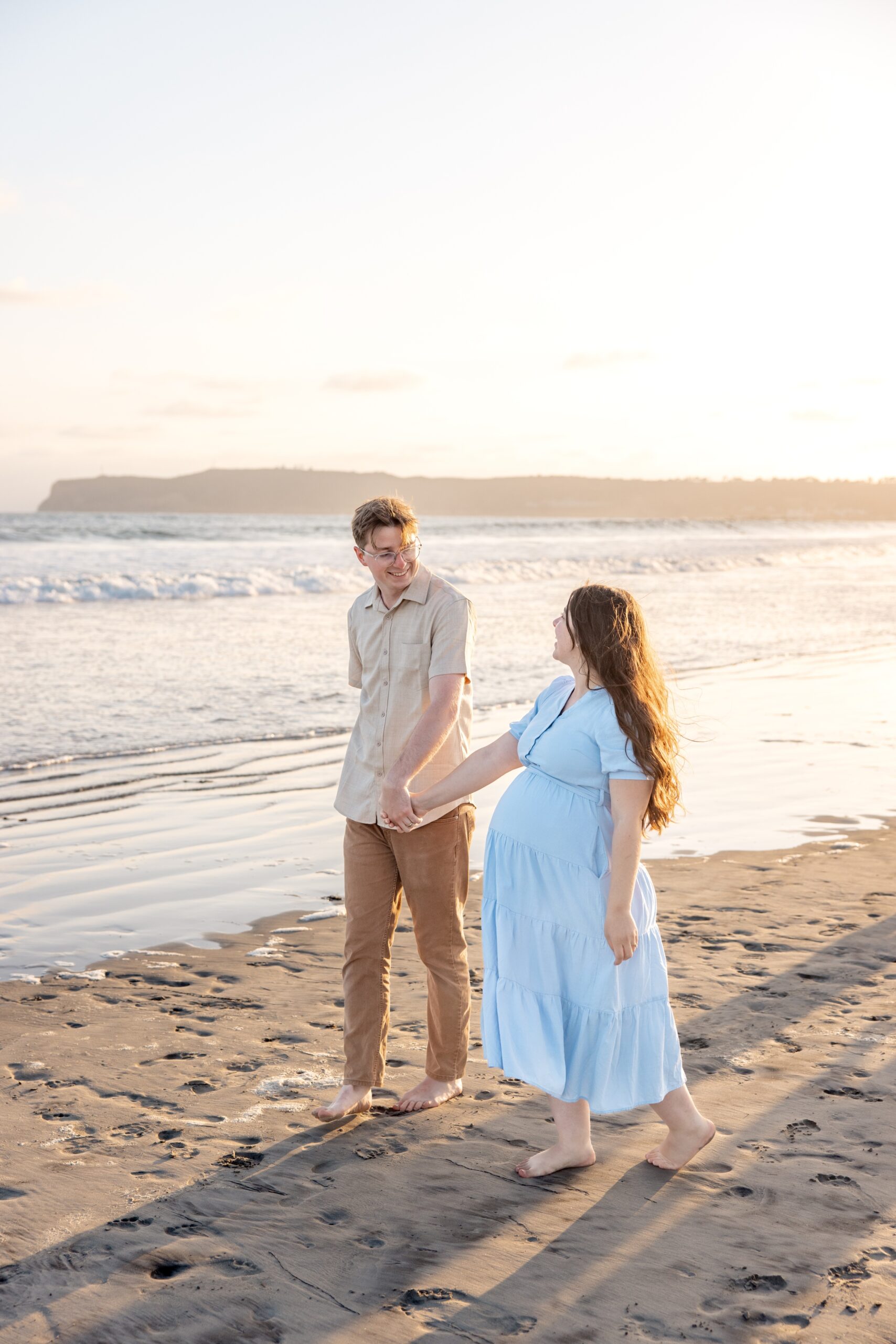 A happy pregnant couple walks on a beach in a blue maternity gown holding hands at sunset after visiting private golf courses in san diego