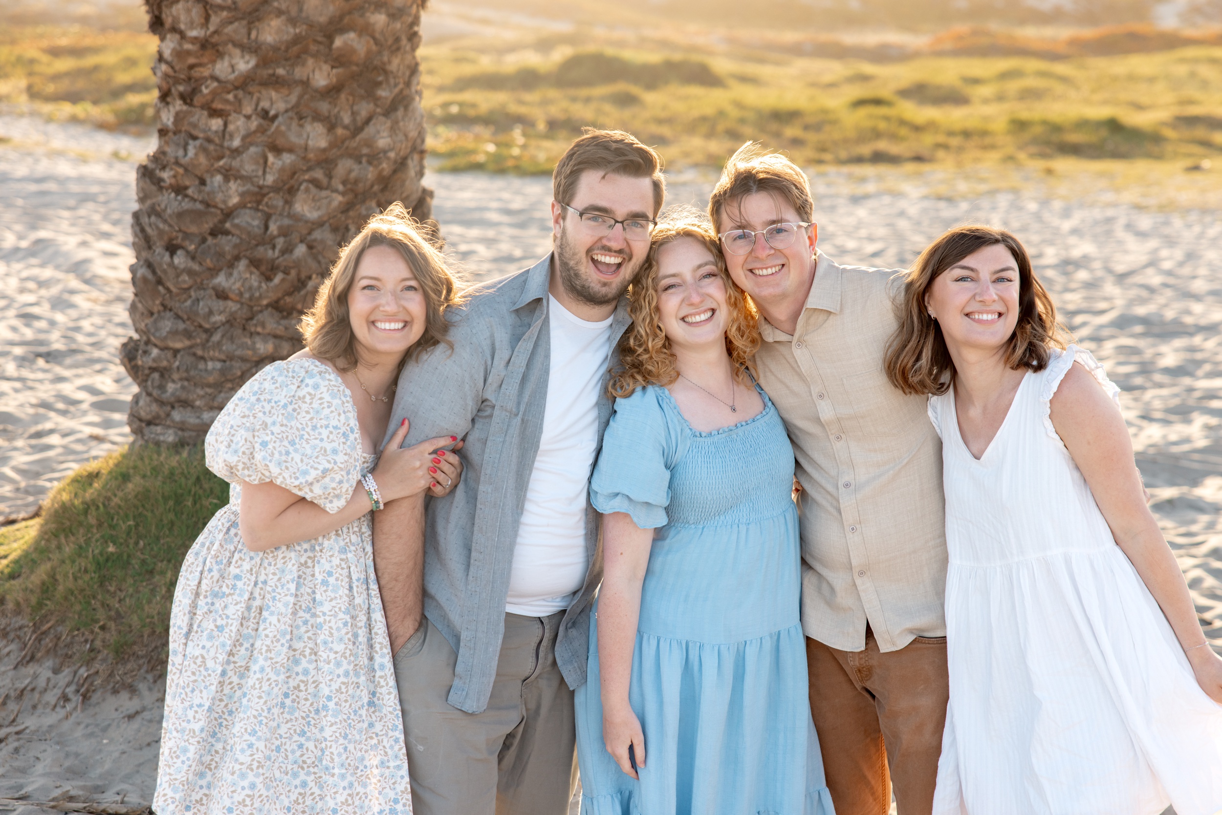 A group of happy siblings stand together having fun under a palm tree at sunset on a beach after visiting private golf courses in san diego