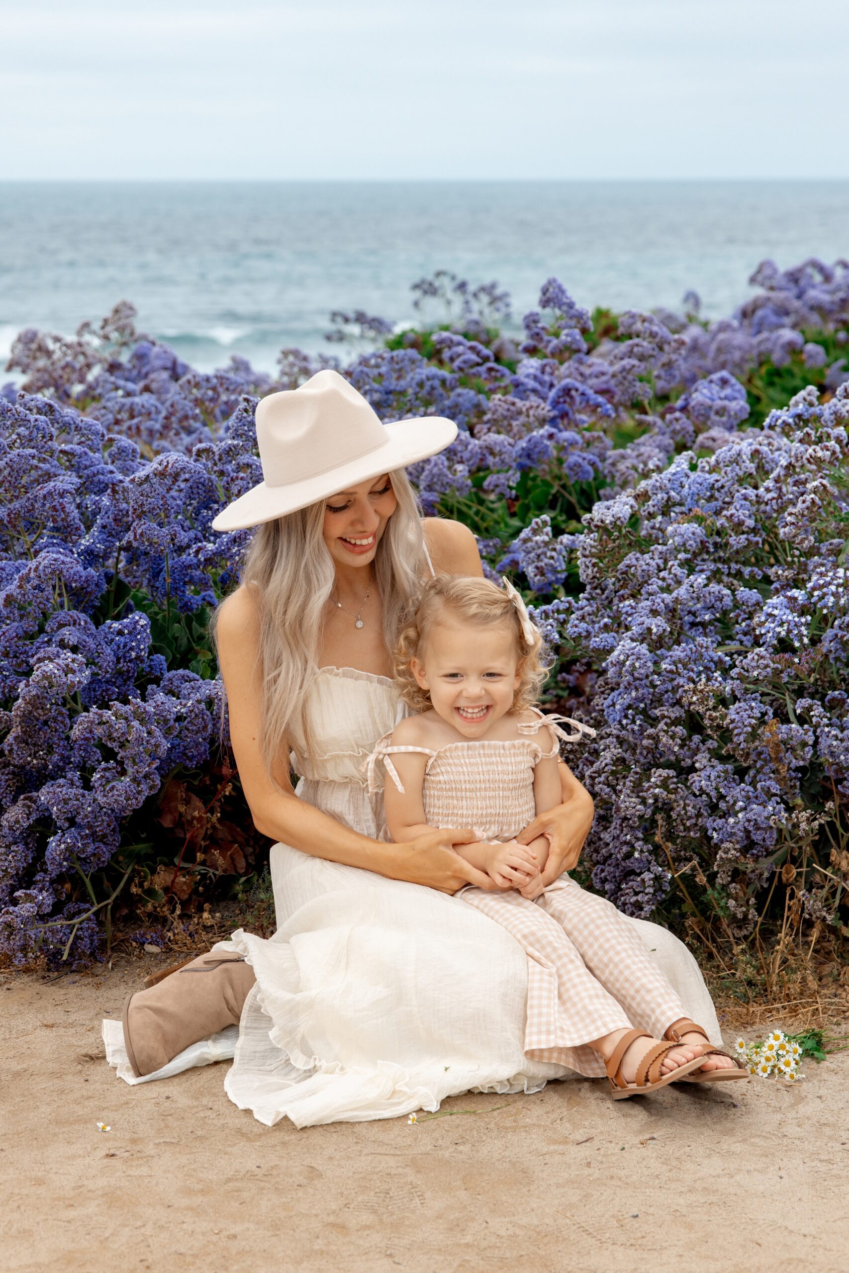 A happy mom in a white dress and large hat sits with her toddler daughter in her lap surrounded by purple wildflowers overlooking the beach