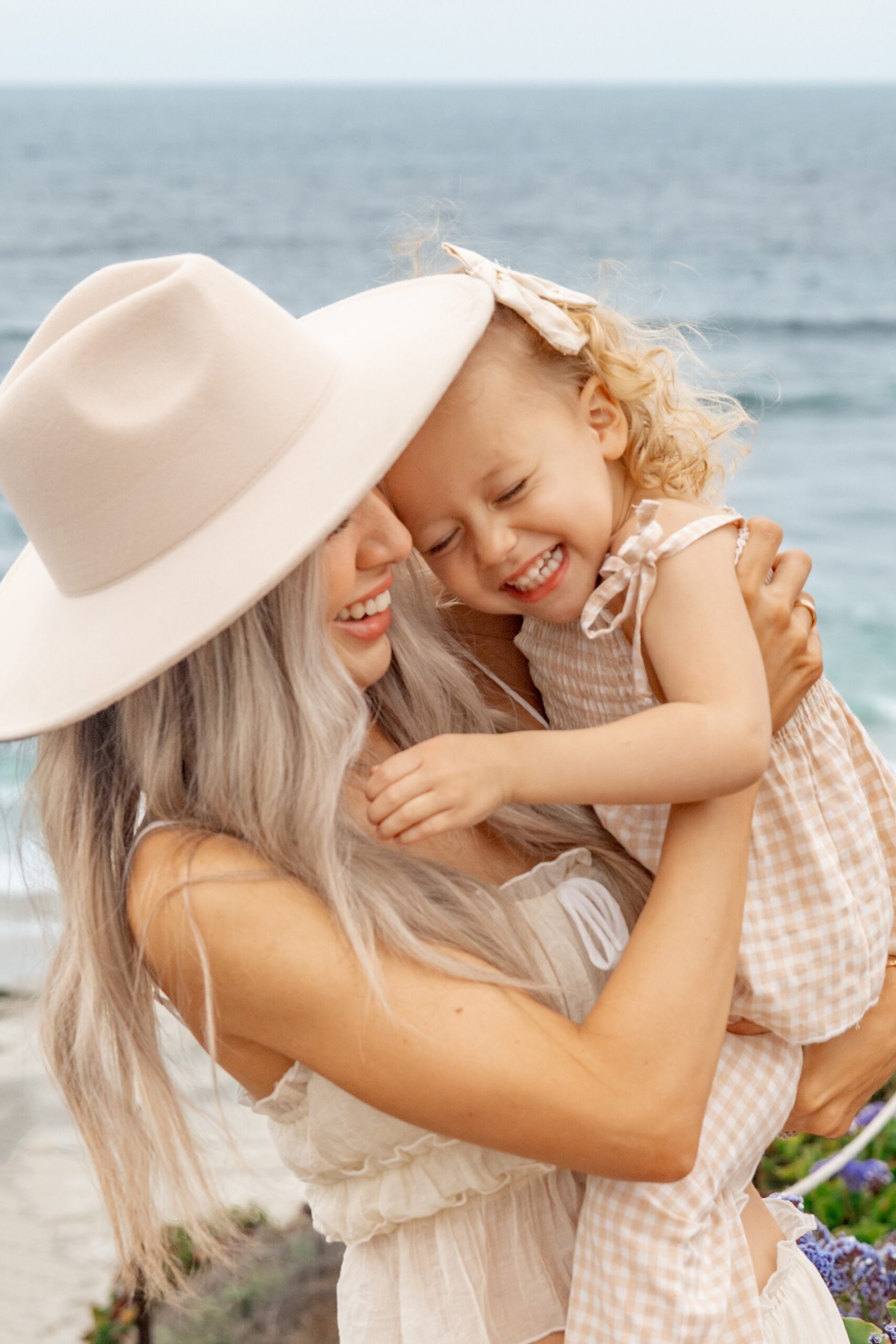 A toddler girl in a cream dress giggles while being lifted by mom on a beach before visiting san diego restaurant week