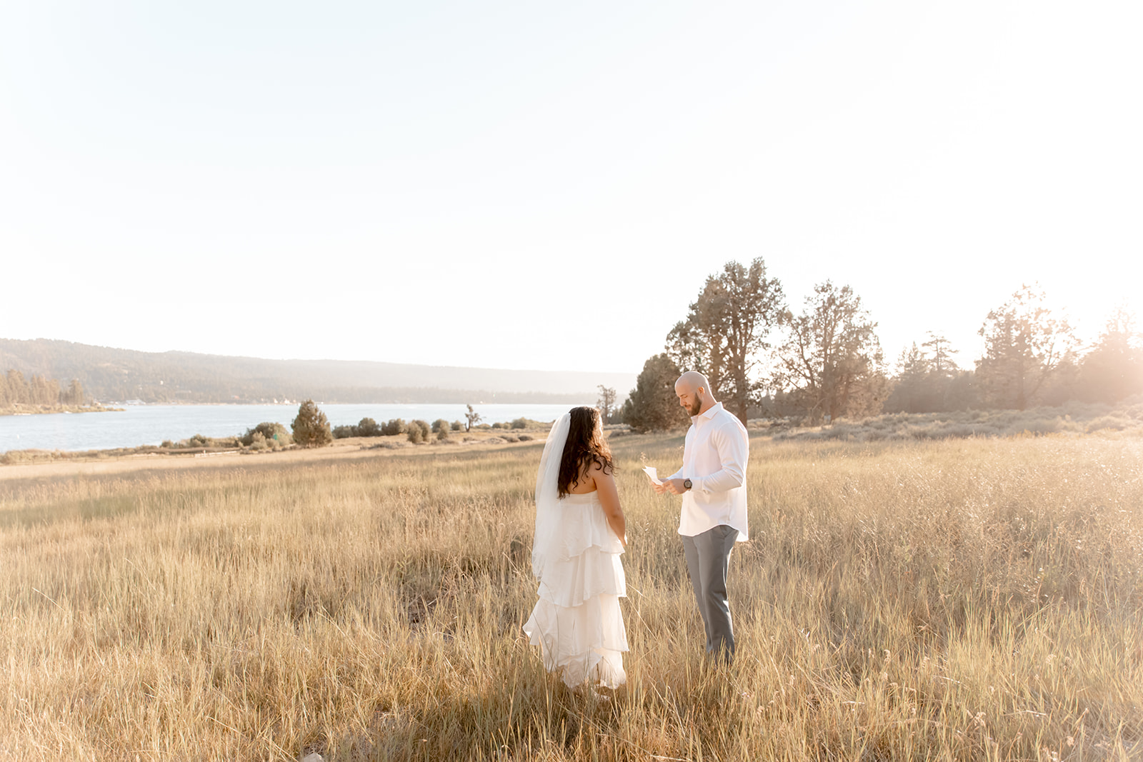 A bride stands in a field of tall golden grass by a large lake as her groom reads a note in a white shirt after finding perfect San Diego wedding Planners