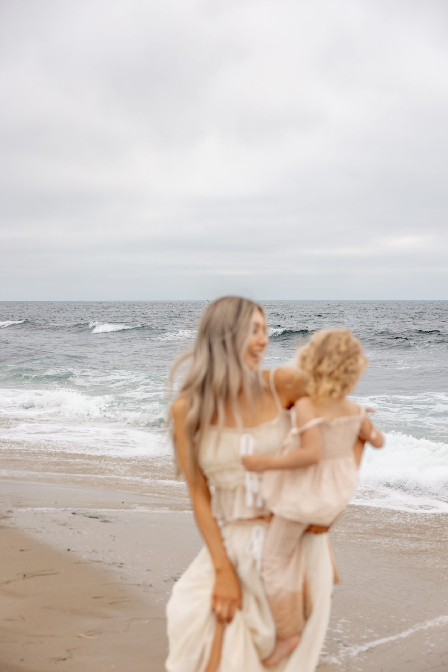 A happy mom dances with her toddler daughter on the beach in a white dress