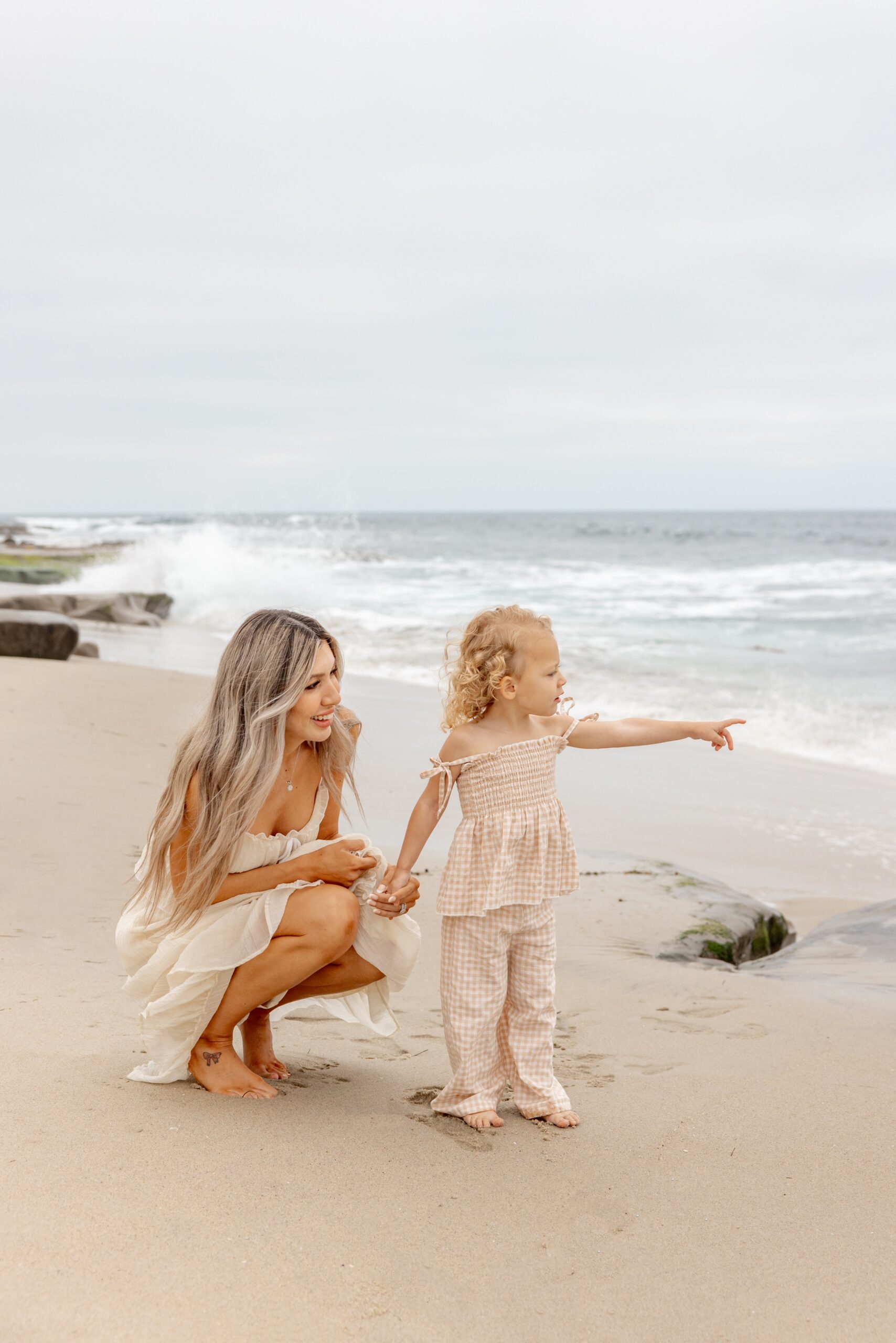 A toddler girl in an orange outfit points to the ocean to show mom something as she kneels behind her holding her hand after visiting the playground san diego