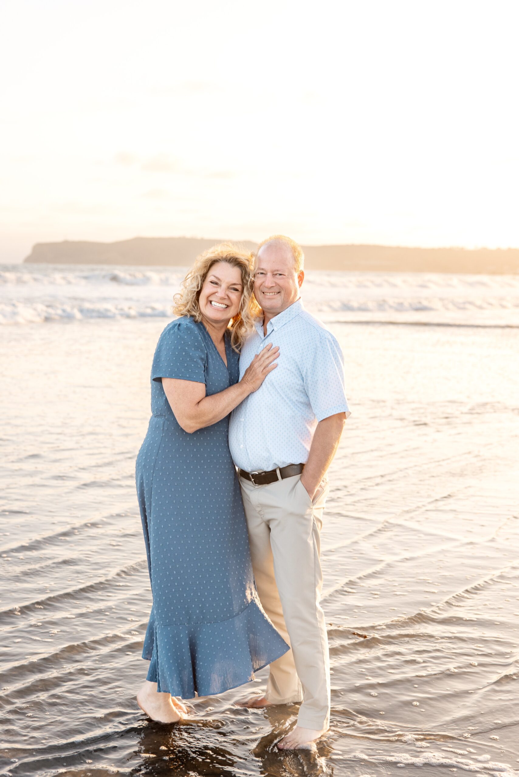 Happy grandparents in blue snuggle on a beach in the shallow water while standing at sunset