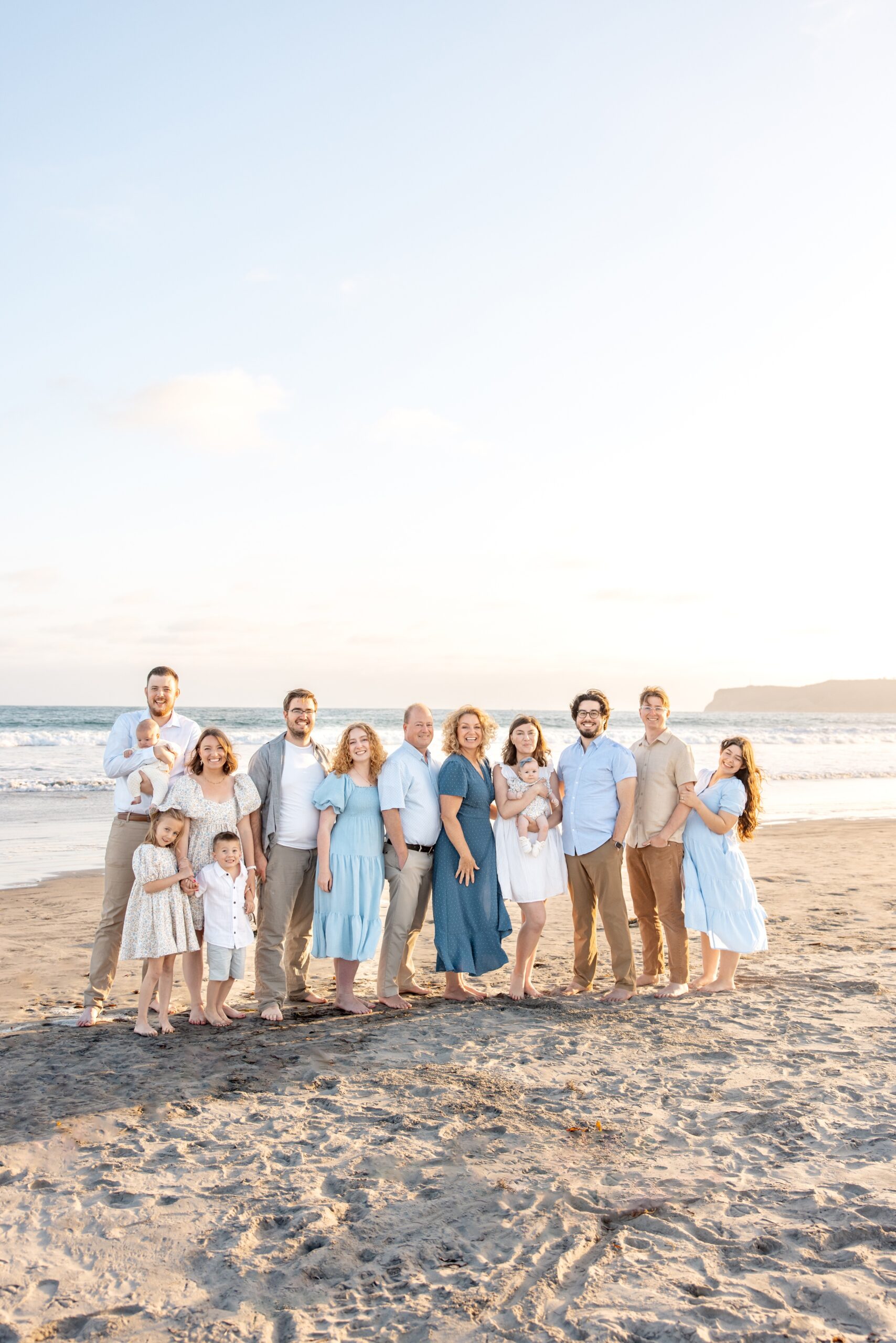 A large extended family smiles while standing together on a beach at sunset before some wine tasting in san diego